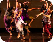 Nine femme dancers curve their arms in luscious extension, facing all directions but connected to each other in warmth. They are caught, in this photo, mid-movement, one knee bent and foot flexed, ready to take the next grounded step. Their skin is of varying brown skin tones with varying hair lengths and textures; their wear silk dupioni costumes (sleeveless tops and pants) in various colorblock shades of fuscia, lime green, lavender, silver, red, and amber. In the foreground on the right, there is a neat stack of short, ceremonial brooms, with silver and blue silk-wrapped handles. The background is a warm brown with soft small "fireflies" of light scattered throughout.