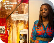 Medium brown skinned woman, blue dress, standing to the right of a large painting with Jim Crow images and the state of alabama painted upon it depicting expressions of capital punishment.