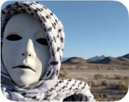 A masked figure with a death-white face, solid black eyes, and a down-turned mouth, wearing a black leather jacket and a keffiyeh (black and white patterned headscarf). The figure stands in front of a desert landscape with blue sky, patchy desert greenery, and distant mountains. Text above the image reads: “DRONE by Andrea Assaf.”