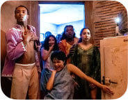 Five dancers pour out of a doorway towards us. To the far left, a medium-dark skinned dancer with short black hair stands with his hands over his chest. He is wearing a pink-lavender open shirt with tan pants. To his right is a medium-light skinned dancer wearing a white, sleeveless shirt and shoulder-length black hair. She is smiling with one hand on their chin. To the right of them stands a dark-skinned dancer with her mouth open in shock. She is wearing a cream and orange top and has her hand stretched out on the arm of a dancer standing in front of her who is wearing a green dress. The dancer in the green dress has a pensive expression. Centered, and in front, is a medium-skinned dancer wearing a blue-grey dress. They have one knee on the ground, the palm of their hand on their chin, and a joyful, mischievous expression on their face. They have their other arm extended grabbing onto a cabinet to their right.
