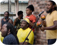 Seven dark skinned men perform music in the back yard of a brick house. They play drums and shakers, and some sing into microphones.
