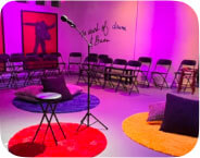 A lone microphone stand is spotlit is in the center of a small room lit with multi-colored lights. Pillows on the floor, colorful carpets and chairs surround the microphone stand.