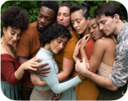 Seven dancers of different races embracing one another in a collective hug with their eyes softly closed.