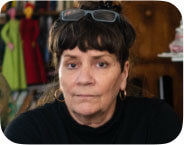 A light skinned mature woman dressed in black is seated looking straight at the camera. Behind her are books and masks and materials for creating puppets. In her hand she holds a small puppet head.