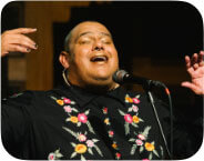A medium-skinned trans femme person with her mouth open in front of a microphone. Her arms and hands are raised at face level. She is wearing a black robe-like dress with embroidered flowers of varying colors.