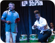 A group of musicians singing and playing instruments on a stage at La Pena Cultural Center.  An Afro-Cuban male drummer playing congas.  A dancer with arms lifted midway and his feet lifted off the ground.  The lead singer, a medium-dark-skinned Cuban man, wears a grey Guayabera and is mid-song, facing the audience and microphone.  Two musicians are playing large chekeres.