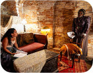 A medium-skinned Black woman is seated, on the left-hand side of the photo,  on a dark red chaise lounge. She is wearing a white corset and a blue dress.  To her right, standing, a medium-dark-skinned woman is looking at her, dressed in a purple nineteenth-century style dress.  The room around them is full of fabric and 19th-century furniture. There is a brick wall behind them, and in front of the chaise lounge a trunk with a white fabric covering it. On the far right side of the photo is a mirror.  A red patterned rug is on the dark wooden floor underneath them.