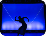 A dancing silhouette in a skirt with arched in an S curve spine and arms against a blue backdrop with a glowing “V” upstage.