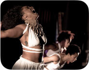 This image consists of four dancers. Chanice Holmes, a medium brown skinned African American female dancer wearing a white wrap crop top, white flowy pants and gold beaded choker with cowrie shells, stands facing to the side with both arms reaching out to the sides, head tilted back and mouth open in a mid-scream. Millenique Brown, a medium brown skinned African American female dancer wearing a white wrap crop top and white pants, stands facing to the side with arms bent in mid-reach up, torso bent forward and legs in mid-step. Catherine Caldwell, a medium brown skinned African American female dancer wearing a white crop tank and white flared pants, stands facing to the side with arms reaching side, torso bent forward and legs in mid-step. Jasmin Simmons-Edmond, a medium brown skinned African American female dancer wearing white wrap crop halter and white flowy pants, stands facing to the side with arms reaching side, torso bent forward and legs in mid-step.