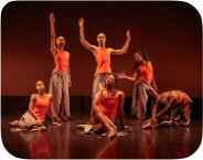 Six Black and Brown of mixed skin tones pose in a group tableaux. They are wearing uniform orange tank tops and grey pants with skirt panels draping below. They are on a stage with a black floor and cyc with faint orange light.