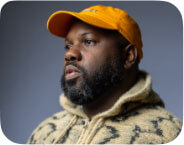 A dark-skin Black man, in a black and tan hoodie and an orange cap. It is a 2024 self-portrait of L. Kasimu Harris, a writer and artist.