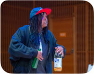 A dark-skinned man with braided dreads,  wearing a dark jacket, a black shirt, and black pants. He also has on a two-tone baseball cap—blue with a red brim. In his left hand, he holds a large glass bottle of Colt 45 malt liquor, and in his right hand, a green glass bottle . Their facial expression is intense and engaged, as if they are mid-speech.