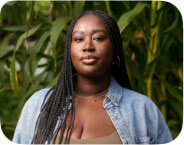 Nana Kumi, a dark-skinned Black woman, stands in a lush community garden on a crisp fall morning, with a strong, rested expression. She wears a green bodysuit and green linen pants, symbolizing her connection to the earth, and a denim shirt honoring the legacy of Black laborers. Her box braids fall over her right shoulder, while the left side is tucked behind her back. One hand rests on her hip, thumb gently tucked into her waistband. She stands grounded and centered in the community garden of a beloved elder, surrounded by plants and the quiet power of ancestral presence.