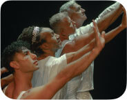 The image shows four dancers in a unified, graceful pose, their arms extended forward in a reaching gesture with flexed hands. Each dressed in various styles of shimmering white costumes, the dancers’ bodies are bathed in soft light that highlights their forms. The front dancer, younger with curly brown hair, gazes upward, while the three elder dancers behind echo the same gesture, their expressions full of intensity and warmth. The dark backdrop amplifies the sense of unity and reverence in this intergenerational moment, evoking themes of strength and connection.