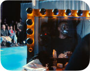 In the foreground on the right, is a large, illuminated dusty dressing room mirror outlined with round black-and-gold bulbs. Reflected in the mirror is a Black male performer, seated wearing a black shirt. His face is painted in black makeup—with exaggerated white paint around the mouth and eyes. He holds a glass in one hand, and his expression appears still and solemn. In front of him on the dressing table, there are a white rag with a red makeup stain, a satin black glove, a clear spray bottle and a sponge soaked with black makeup. Behind him, the mirror reflects a hanging white curtain and white dress shirt and grey jacket hanging from a hook. To the left, in the background, sits an entirely light- skinned audience in a dimly lit space. The crowd is mixed in gender and dress, with people sitting closely in folding chairs. They face the stage, looking towards the performer. Their expressions range from neutral to serious. The walls behind them are dark with exposed red brick pillars, and overhead lights cast a cool glow on the bright blue stage floor, which is outlined with a few white stars.