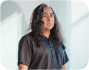 Composer Raven Chacon stands upright in the center of a well-lit room, wearing a black short-sleeved button down and black pants. He is standing behind a folding table on which sits an audio mixing console and a pair of black headphones. He is staring into the camera, with light from the window framing his figure on the wall behind him.