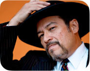 A medium skinned Chicano man dressed in a pin-striped suit, with a white shirt and a multi-colored tie that features round peace symbols. His facial features include a moustache that connects to a goatee. His left hand holds his left lapel, as his right hand reaches for his black, wide-brimmed pork pie Tando (hat), as if about to remove it from his head. The background is covered in a brown/orange tone.