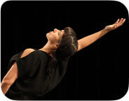 Against a jet black background a Native woman with medium brown skin, dark brown straight hair in a single loose braid and regal facial expression arches back in a dancerly position with her right arm raised high, looking up. She wears a black outfit with a sleeveless top.