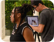 A brown-skinned woman with hair in locs and camera in hand looking at Da Brat and Judy as she gives direction on how to pose them.