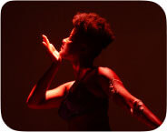 A brown-skinned woman mid-movement dancing. Her right arm is bent at the elbow, heel of palm cupping her chin as she looks to the right. Her left arm is outstretched, and white wax can be seen clinging to her skin on that arm. She is wearing purple rhinestoned bra, garters, and thong, which are shadowed in the spotlight.