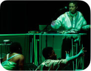 Five black performers awash in green light, gather around two tables set up with sound tech. Jeremy and Holland stand behind the tables as they adjust sound boards and computers; keyon, Katrina and jess rest on the ground, mid-movement.
