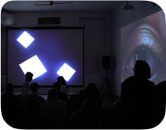 A dimly lit room is filled with an audience seated on the floor, facing three large projections that span the front and side walls. The central projection shows animated white squares of varying sizes floating against a black background. On the left and right walls, close-up videos of a brown-skinned, South Asian person's eye dominate the space, each with a glistening reflection of stars or lights in the pupil. In front of the central screen, silhouetted audience members are visible, with a few standing or moving about. Among them, one person with medium-dark skin and short curly hair appears to be operating audiovisual equipment on a table. Two ceiling-mounted projectors and large speakers are visible above and around the room. The atmosphere illuminates the activation of an immersive audiovisual performance and video installation.