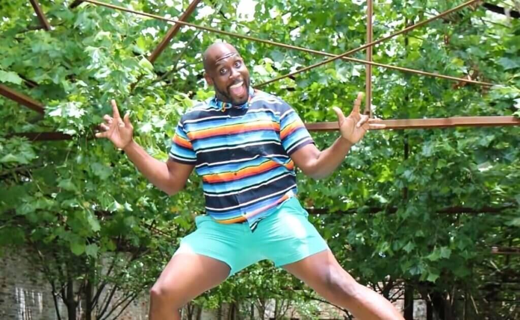 Addam Garrett, a Black man with a bald head, mid-jump, smiling widely and pointing his fingers upward. He is wearing a colorful, striped shirt and turquoise shorts. The background features rusted rafter beams from what was once a warehouse space, now open to the elements, with lush green foliage growing around the area.