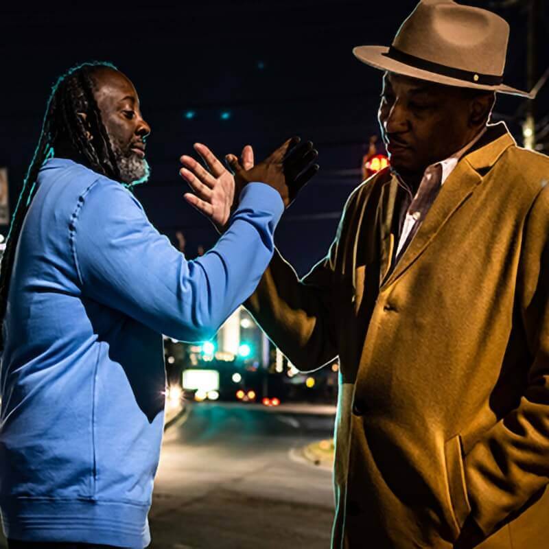 This image shows two men, identified as artists Bluz (on the left) and Dasan Ahanu (on the right), sharing a handshake or hand gesture on a city street at night. Bluz is wearing a light blue sweatshirt, while Dasan Ahanu is dressed in a brown coat and a stylish hat. The scene is set against a dimly lit urban backdrop, with streetlights and traffic lights in the distance, adding a moody and atmospheric feel to the image. The interaction between the two men suggests a moment of camaraderie or mutual respect.