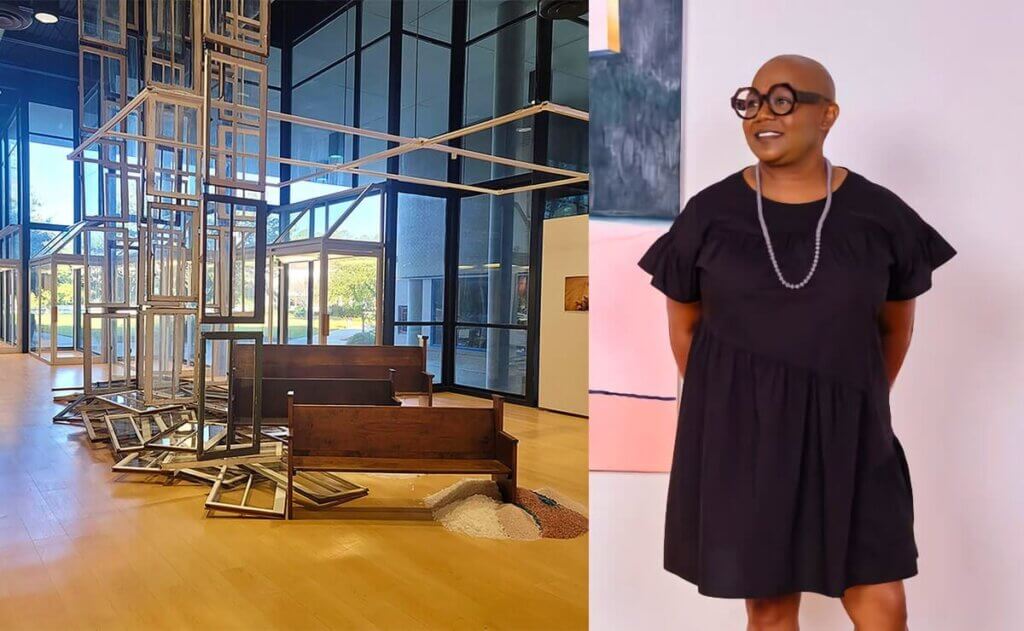 This image is split into two sections. On the left is an art installation inside a large, modern building with glass walls. The installation consists of wooden church pews, scattered window frames, and a tall, geometric structure made of more window frames, creating an abstract and contemplative scene. On the right side of the image is a portrait of Michaela Pilar Brown, who is standing against a neutral background, smiling and dressed in a simple black dress with a beaded necklace and large, round glasses. The contrast between the dynamic installation and her poised stance highlights her as an artist or curator associated with the installation.