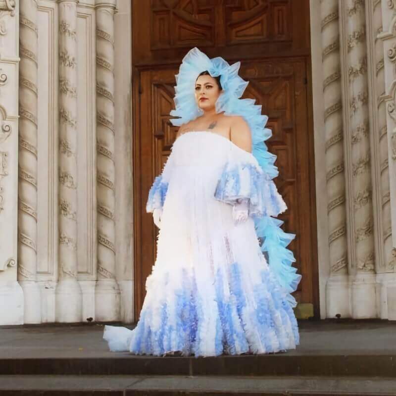 This image shows San Cha standing on the steps of an ornate building, possibly a church or historic structure, wearing a dramatic and elaborate gown. The dress is predominantly white with soft blue accents and features voluminous, ruffled sleeves and a matching headpiece that frames her face. The outfit has a regal and ethereal quality, complemented by the grand architectural backdrop. San Cha's pose is confident and commanding, highlighting her presence and the intricate details of the gown.