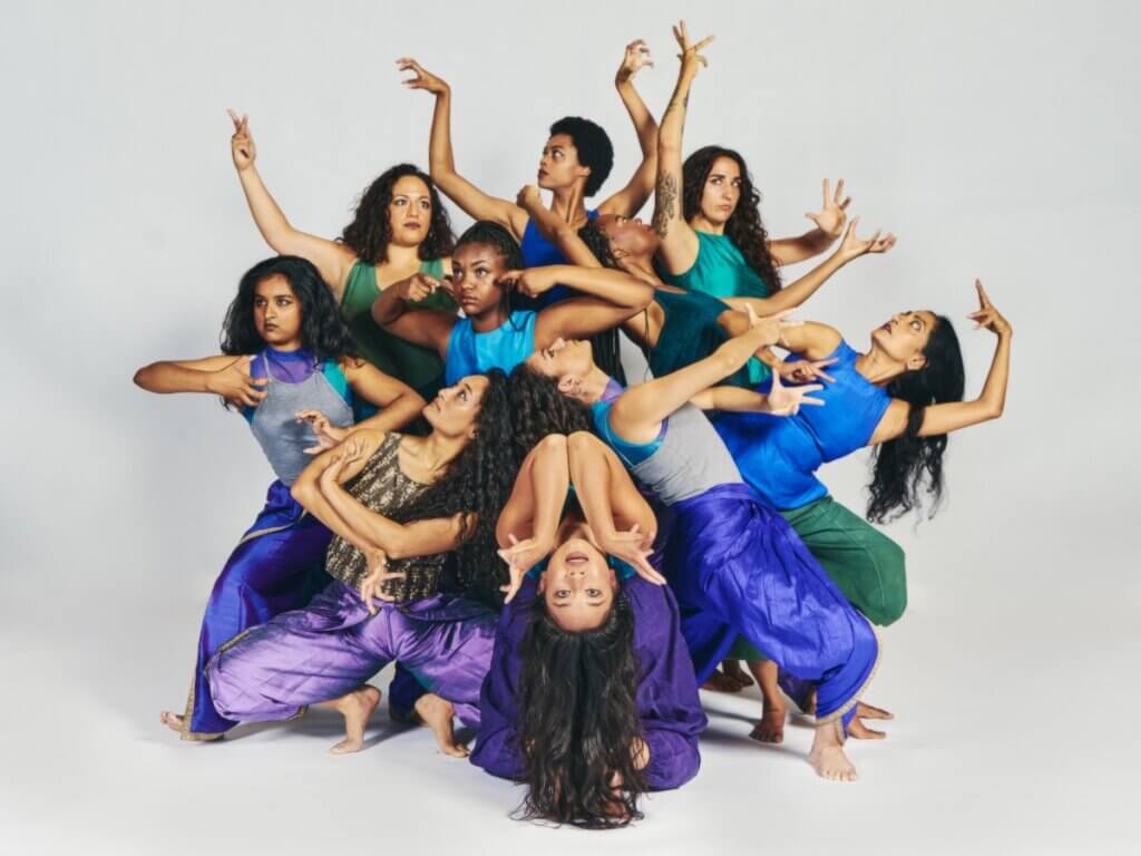 A group of women and femmes pose in a circle, each striking a position of specificity and power, facing in all directions, most away from the camera. The woman at the bottom of the circle poses in a backbend facing the camera, her arms framing her upside-down face. All performers are BIPoC and each woman/femme is of different skin tone, hair texture, and body shape. They wear close-fitting silk shirts in blue and green jewel tones and loose silk pants in blue, green, and purple jewel tones. 