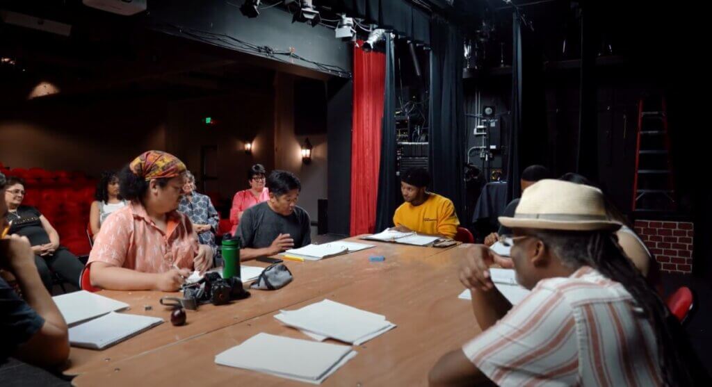 A table of Black and Japanese performers, a playwright, and director all sit around a brown rectangular table on the stage of a dark theater, printed out scripts are in front of each of them as they read the play together for the first time. They are all focused on the current actor reading lines at the center of the photo. The viewer is positioned as if sitting at this table. In the background, four audience members look on from further downstage. 