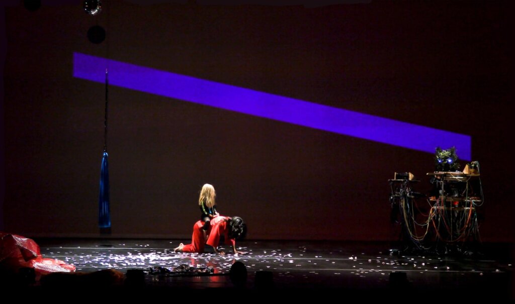 A blue compression swing hangs from the ceiling upstage, stage right. A blond-haired child sits on the back of a dark-haired woman wearing all red. She is crawling. There are pink petals beneath her. A person wearing a tree mask is behind a table of synthesizers, stage left. There is a projection of a large purple bar on the scrim behind the performers.
