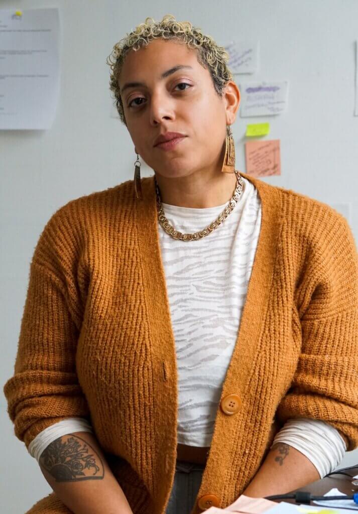 A medium brown skinned person sits behind a desk covered in different colored post-it notes facing the camera. With short-cropped hair and chunky gold jewelry, they face the camera wearing a pumpkin-colored cardigan, a white shirt, and light blue ripped jeans.