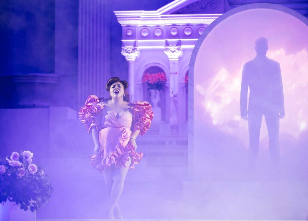 Latinx performer San Cha, a medium-skinned woman, standing triumphantly in a knee-length, peach-colored dress adorned with ruffled shoulders and skirt. She poses gracefully in front of a marble arch within a cathedral adorned with roses, where the projection of a foreboding masculine figure in a suit adds an intriguing contrast to the scene, all set amidst dense fog.