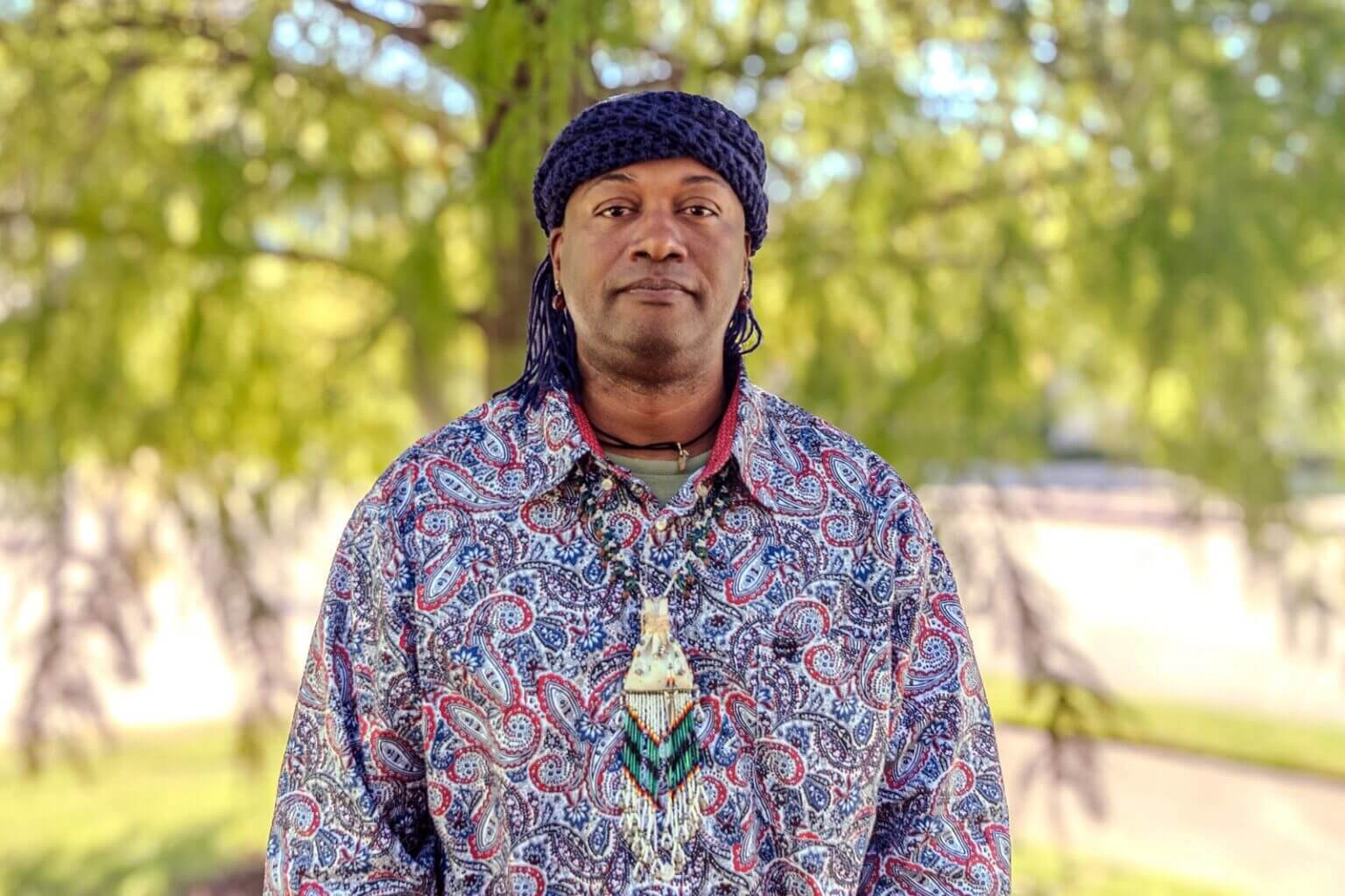 A man, Nant'a Cougar Goodbear, wearing a patterned shirt, a headscarf, and a beaded necklace, stands outdoors with trees in the background.