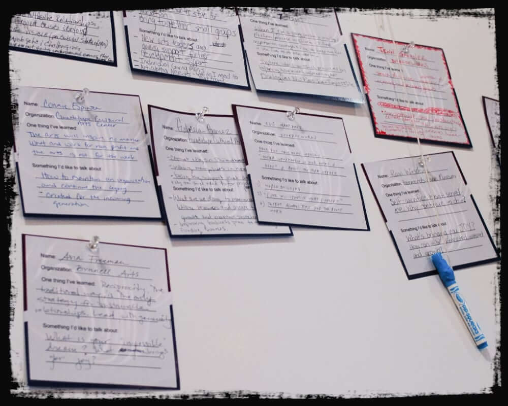 Photo of a display wall featuring multiple handwritten “dance cards,” each listing participants’ names, organizations, something they’ve learned, and topics they’d like to discuss—ranging from funding and leadership to community collaboration. Cards are pinned with clips, and a blue marker dangles on a string, suggesting interactive use.