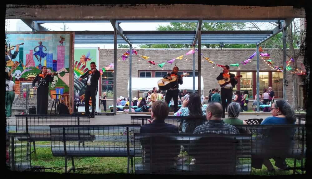 A lively outdoor performance features a mariachi band playing on a small stage, with musicians dressed in traditional black charro suits. Colorful papel picado decorations hang above the stage, and an audience sits on metal benches enjoying the show. A mural and festive community gathering provide a vibrant backdrop, suggesting a cultural celebration or local event.
