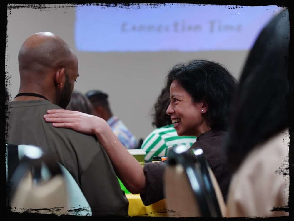 A smiling woman places a comforting hand on a man’s shoulder during a group activity, both seated among others in what appears to be a workshop or seminar. In the background, a projection screen displays the words “Connection Time,” reinforcing the theme of interpersonal bonding.