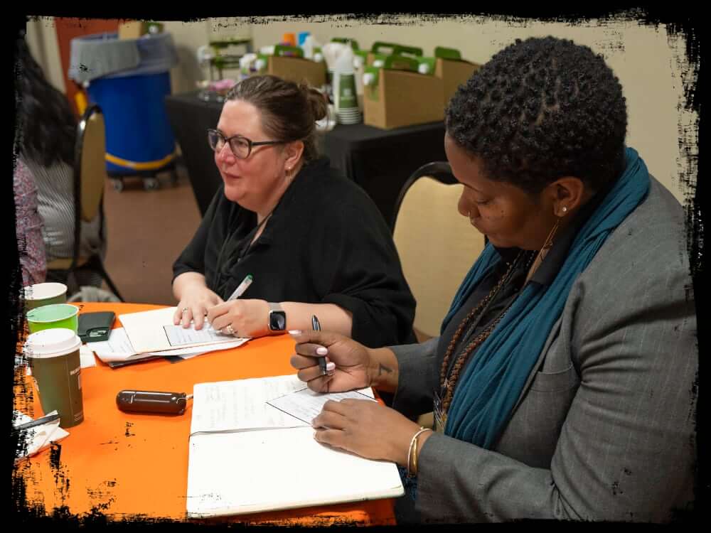 Two women are seated at a table, actively engaged in taking notes during a meeting or workshop. The woman on the left wears glasses and a smartwatch, smiling while holding a pen, and the woman on the right is focused on writing in a notebook, dressed in a blazer and teal scarf. The table has papers, notebooks, coffee cups, and a phone, with a background showing catering supplies and cardboard boxes.
