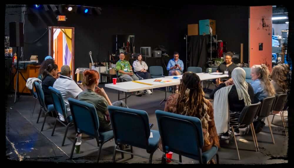 A diverse group of people sit in a circle of chairs around two rectangular tables in a dark, studio-like room, engaged in a group discussion or workshop. The space includes backstage equipment, exposed storage, and a colorful curtain at the open exit door, adding a creative, behind-the-scenes vibe to the gathering.