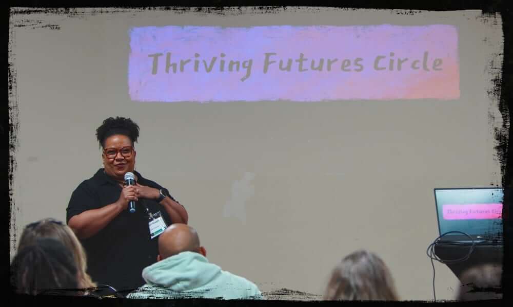 A speaker stands at the front of a room holding a microphone, engaging an audience during a session titled “Thriving Futures Circle,” as shown on the projected slide and laptop screen. The setting suggests an inclusive, interactive workshop or discussion focused on forward-thinking community or arts topics.