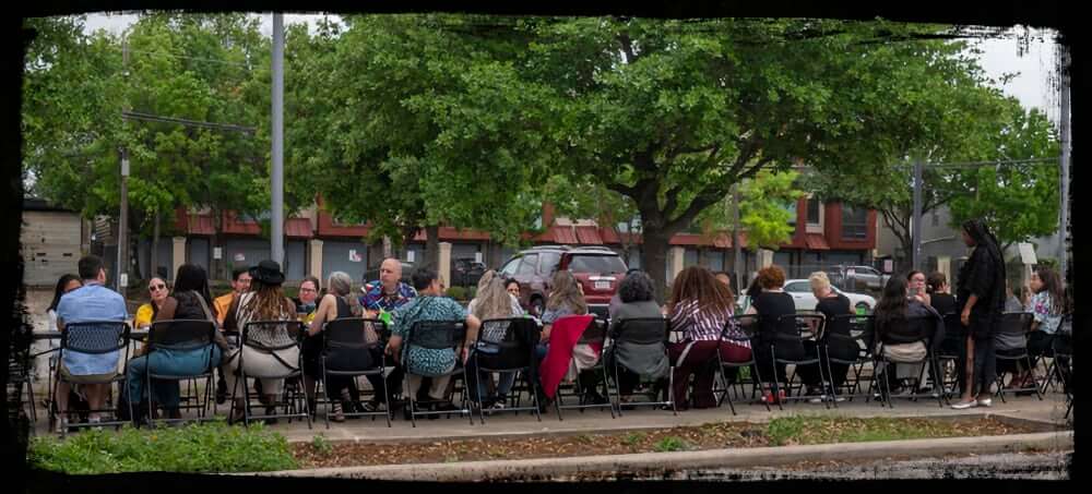 A large group of people sit together at a long table set up outdoors along a city street, enjoying a communal meal under the shade of leafy trees. The relaxed, informal gathering suggests a community dinner or networking event, with conversation and connection at its core.