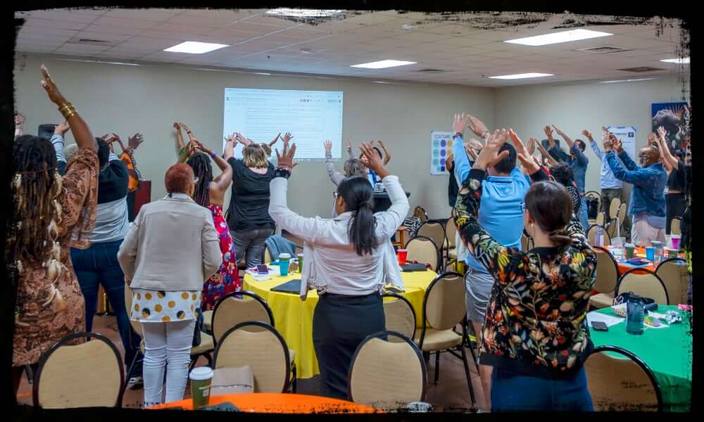 A large group of workshop participants stand beside round tables covered in colorful tablecloths, raising their arms in unison during a group exercise or energizer. The scene suggests a lively and inclusive atmosphere, with a presentation projected on the wall behind them.