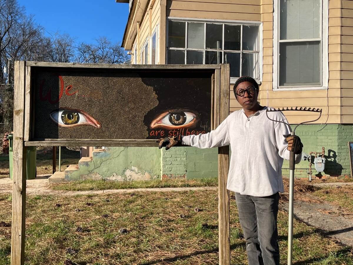 The Black artist Jahni Moore stands outside next to a hand-painted sign that displays two large eyes and the words, "We are still here." He is wearing faded jeans, a loose white shirt, glasses, and gardening gloves, and he is holding a rake. The sign is approximately five feet wide by two feet tall, and it's raised about four feet off the ground by two wooden posts. Behind him and the sign is a partial view of the Drake House, with yellow siding and a concrete-and-brick foundation that's painted a pale green. The sky is a clear blue, and the trees in the distance are bare, suggesting the photo was taken in late winter or early spring. 