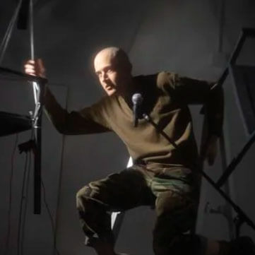 Joshua Kohl performs in Boy Mother Faceless Bloom crouched on a dimly lit stage while gripping a vertical pole and leaning toward a microphone. Dramatic side lighting highlights his shaved head and tense posture as he appears mid monologue creating an intense and intimate theatrical moment.
