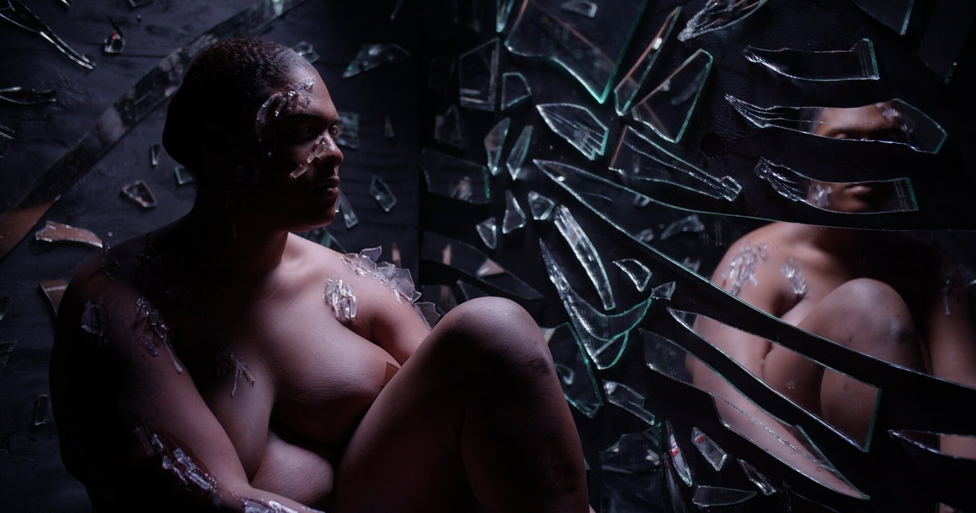 Still from the documentary "Queen of the Pen" showing a Black trans woman sitting nude, with one knee drawn up to her chest, in a dark space next to a wall covered in large shards of mirrored glass. Pieces of irregularly shaped clear acrylic are affixed to her face, shoulders, breasts, and arms to suggest embedded shards of glass.