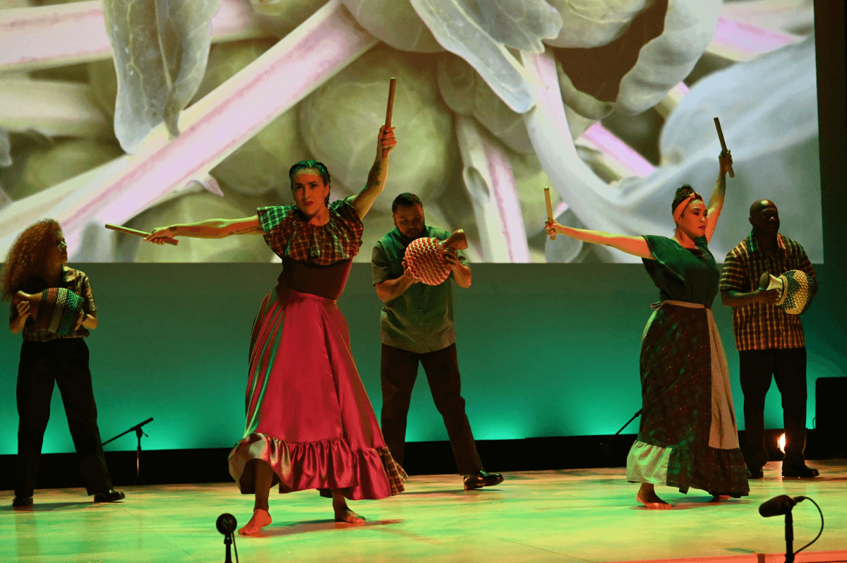 La Mezcla dancers and musicians perform a piece titled “Sembrando.” Dancers are wearing long, flowing skirts and a combination of purple and green tops while holding sticks. 3 musicians play chekeres (hollowed out gourds wrapped in beads) behind them and wear plain black pants and checkered shirts. A close up shot of brussel sprouts is projected behind the performers as the stage is flooded in green light.