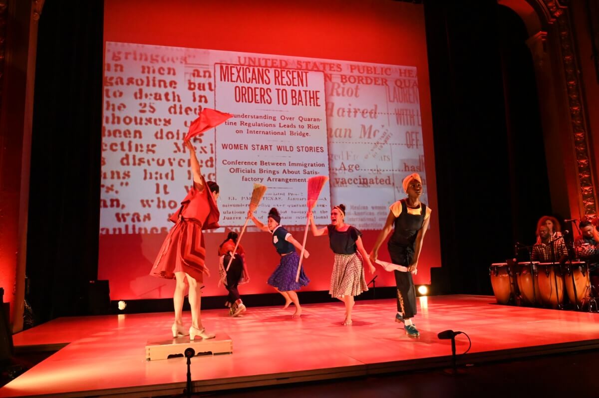 La Mezcla dancers perform a piece paying homage to the bath riots along the US/Juarez border in 1917. Dancers perform in front of an archival newspaper headline that reads, “Mexicans Resent Orders to Bathe '' and “Women Start Wild Stories' ' while the stage is flooded with red lighting.