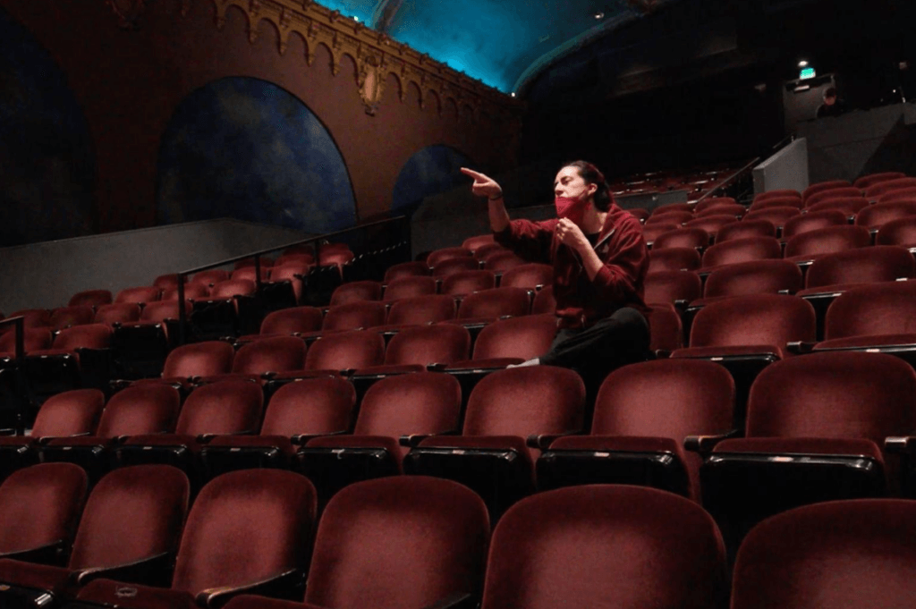 Executive Artistic Director, Vanessa Sanchez, gives dancers notes from the audience. Vanessa is seated in the theater, surrounded by empty seats, and is pointing towards the stage.